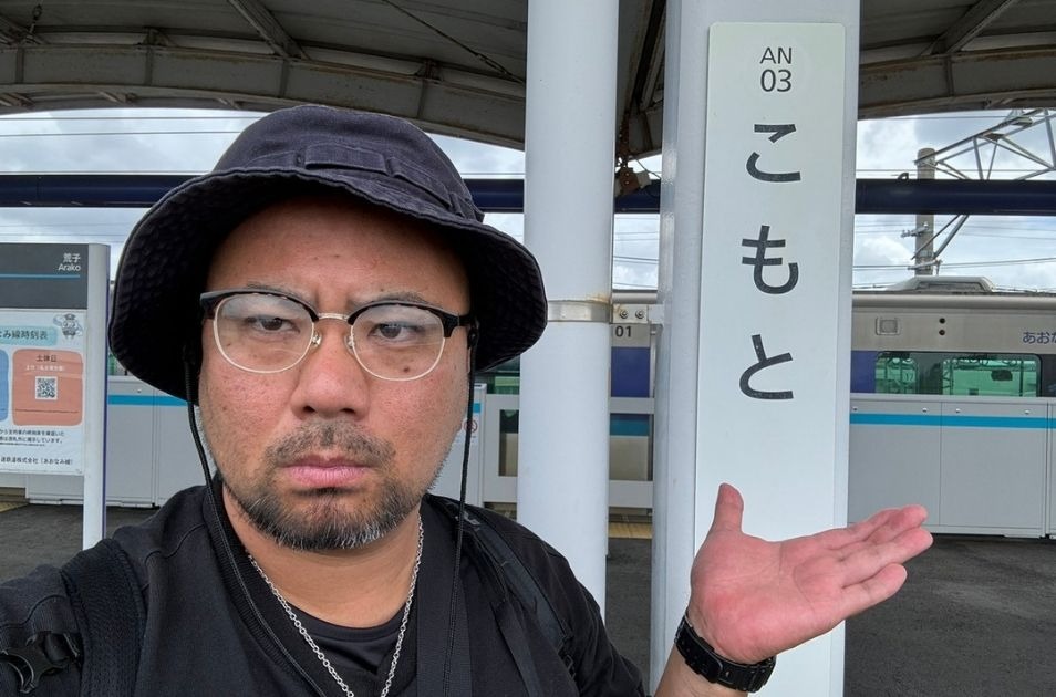 名古屋臨海高速鉄道あおなみ線の「こもと（小本）」駅のホームで、ご自身の名字と同じ駅名標を手のひらで示しながら、バケットハット姿で自撮りをする小元佳祐さんの写真。