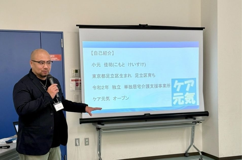 プロジェクタースクリーンの前に立ち、マイクを持ってプレゼンテーションを行う小元佳祐さん。スクリーンには自己紹介として「小元 佳祐（こもと けいすけ）」「東京都足立区生まれ 足立区育ち」「令和2年 独立 単独居宅介護支援事業所 ケア元気 オープン」という経歴と、「ケア元気」のロゴが映し出されている。