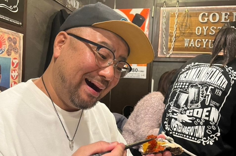 飲食店で、イクラが乗った牡蠣を箸で持ち上げながら嬉しそうな笑顔を浮かべる小元佳祐さんの写真。