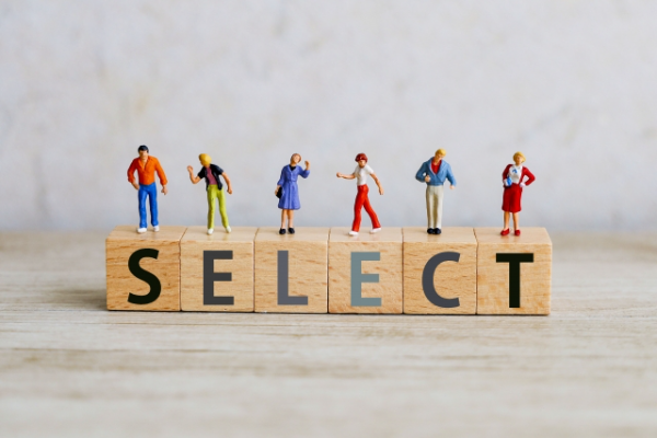 『SELECT』と書かれた木製のブロックの上に、様々な服装の男女の小さなミニチュア人形が立っている写真。