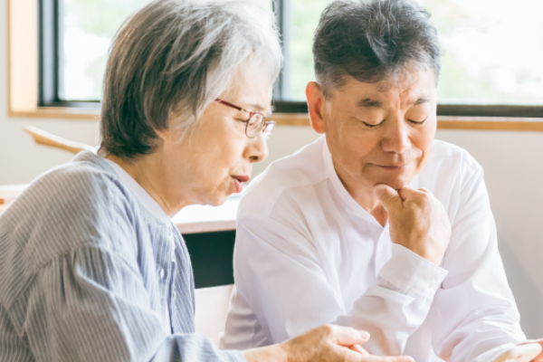 明るい窓辺で、シニア夫婦が一緒にスマートフォンを見つめ、真剣な表情で話し合っている写真。