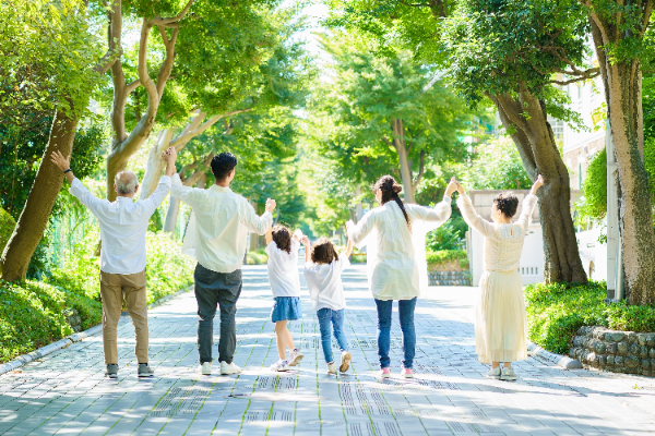 緑豊かな並木道で、シニア夫婦、若い夫婦、2人の子供の3世代家族が手をつなぎ、両手を高く上げて後ろ姿で並んで歩いている写真。