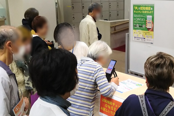『「声」で認知機能をチェックしてみませんか?』というポスターが貼られた会場で、シニアの方々がスタッフの説明を聞きながら、スマートフォンでアプリを体験している様子。