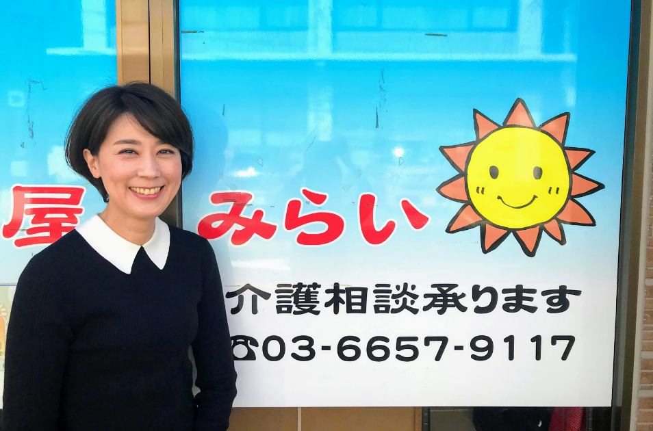『介護相談承ります』と書かれ、笑顔の太陽のイラストが描かれた事業所(みらい)の青い看板の前に立つ、産業ケアマネジャー(服部さん)の写真。