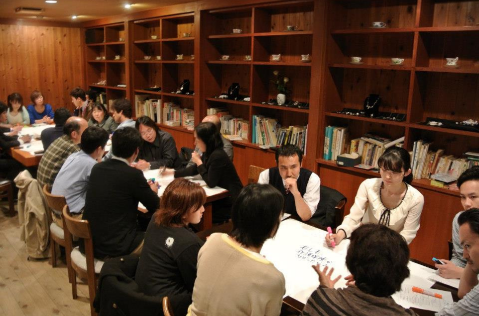 カップが並ぶ棚がある木目調の落ち着いた部屋で、複数のテーブルに分かれた参加者が模造紙を囲み、真剣に話し合っている様子。