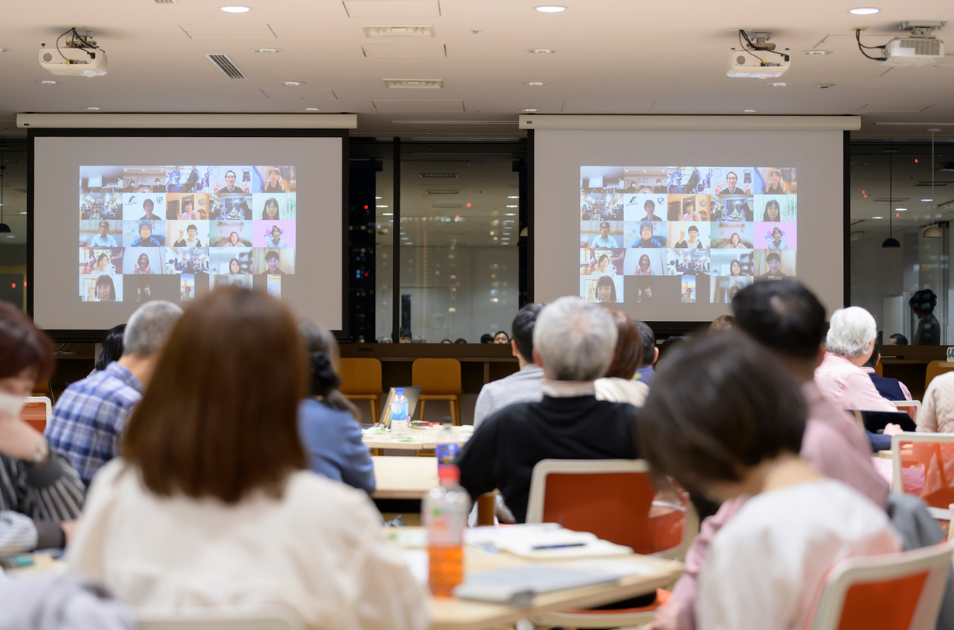 会場の参加者たちが、前方のスクリーンに映し出された多数のオンライン参加者の映像を見ている様子。