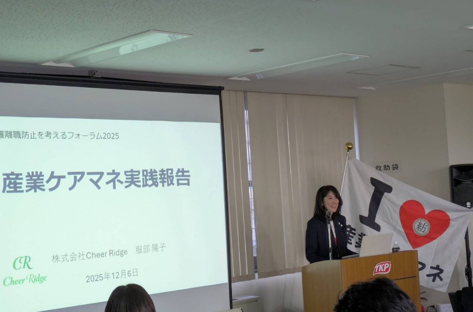 『産業ケアマネ実践報告』と映し出されたスライドスクリーンの横で、TKPと書かれた演台に立ち、マイクに向かって笑顔で講演を行っている服部さんの写真。背後には『I ♡ 産業ケアマネ』と書かれた旗が飾られている。