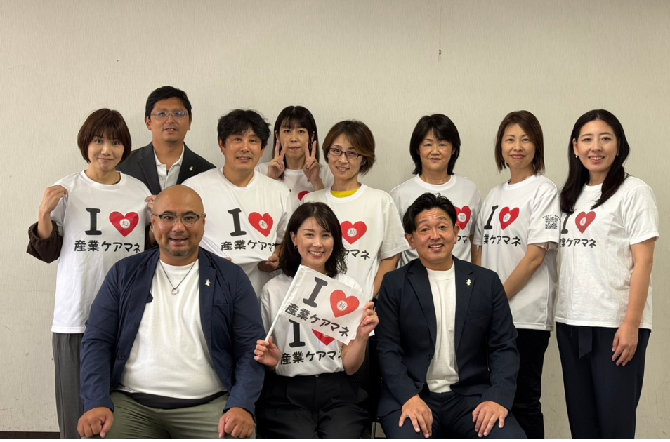 『I ♡ 産業ケアマネ』とプリントされたお揃いのTシャツを着た男女11人のグループが、笑顔で集まっている集合写真。前列中央の服部さんは同じデザインの小さな旗を持っている。