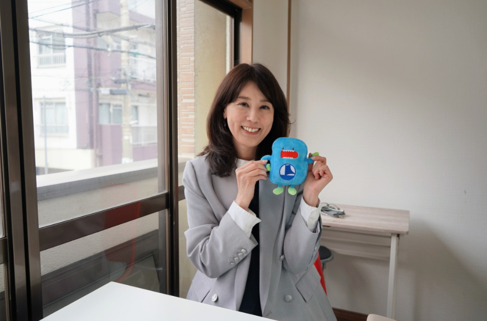 明るい窓辺の部屋で、グレーのジャケットを着た服部さんが、青いキャラクターのぬいぐるみを両手で持ち、リラックスした笑顔を見せている写真。