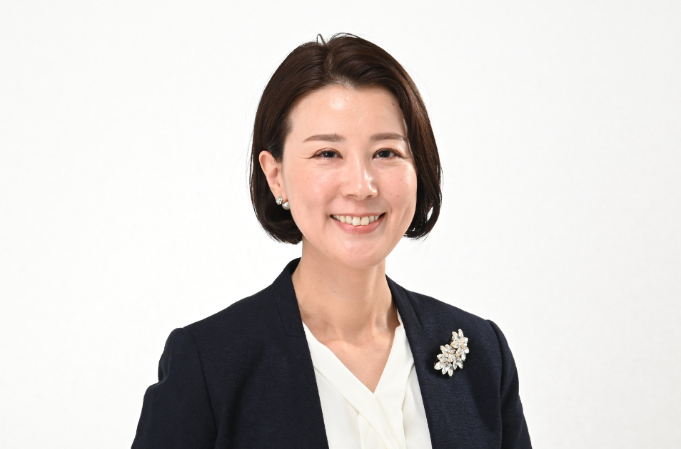 白い背景で、ネイビーのジャケットに白いブラウス、花のモチーフのブローチを身につけた服部さん(産業ケアマネジャー)が、明るく優しく微笑んでいる上半身のプロフィール写真。