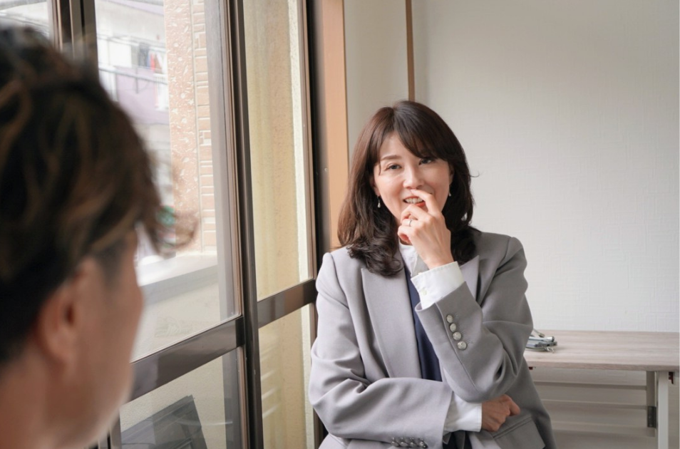 明るい窓辺で、グレーのジャケットを着た服部さん(産業ケアマネジャー)が、インタビュアーの言葉に耳を傾けながら、口元に手を当てて和やかに微笑んでいる写真。