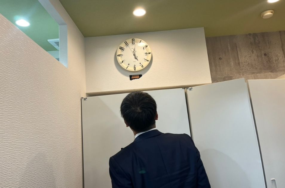 オフィス内で壁掛け時計を見上げるスーツ姿の山梨義正氏の後ろ姿。