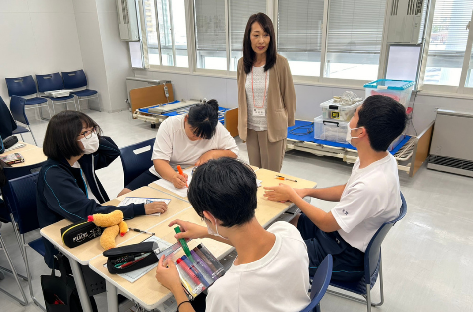 介護用ベッドが置かれた実習室のような場所で、グループワークに取り組む学生たちを温かい眼差しで見守り、声をかけている古池さんの写真。
