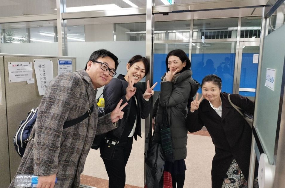 施設内の通路で、女性3名と一緒に笑顔でピースサインをして写真に写る山梨義正氏。