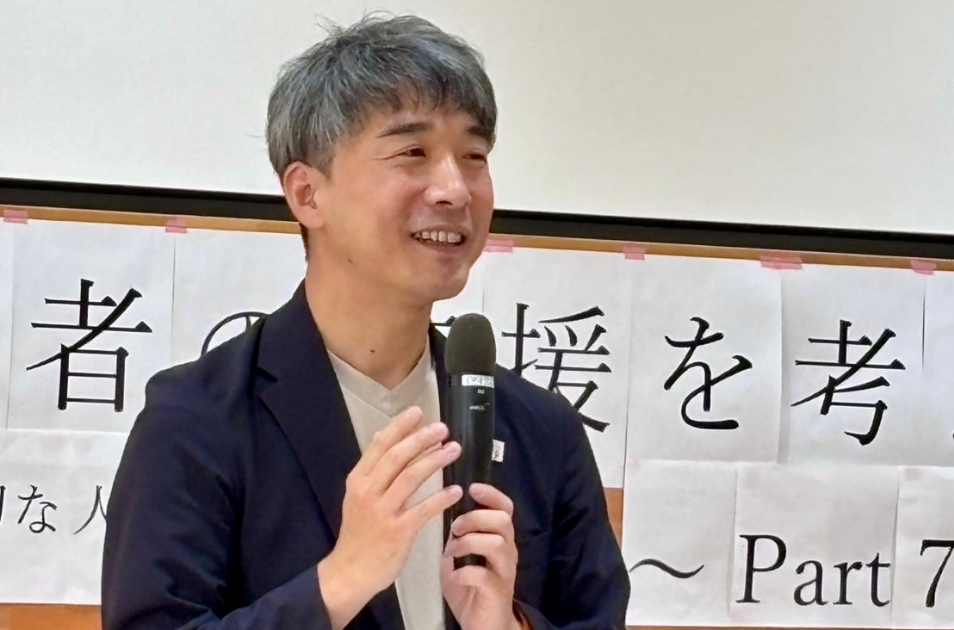 マイクを両手で持ち、笑顔で講演を行っている川上洋平さんのバストアップ写真。背景には『〜支援を考え〜』と書かれた手書きの横断幕の一部が見える。