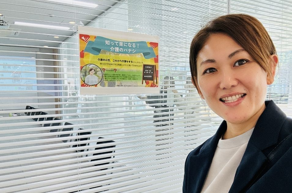 ブラインドに貼られた『知って楽になる!介護のハナシ』という企業向け介護相談窓口の案内ポスターの前で、笑顔で自撮りをする進絵美さんの写真。