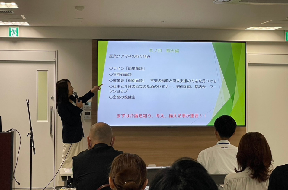 企業向けセミナーの会場で、スクリーンに映し出された『産業ケアマネの取り組み』に関するスライドを指差しながら解説をしている古池さんの写真。