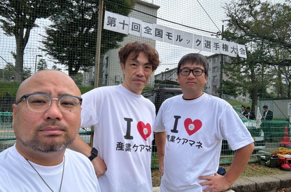 「第十回全国モルック選手権大会」の会場にて、「I LOVE 産業ケアマネ」と書かれたお揃いのTシャツを着て写真に納まる山梨義正氏ら3名の男性。