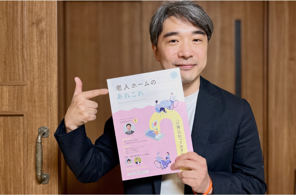 『老人ホームのあれこれ』というタイトルの介護情報誌を手に持ち、指を差して笑顔を見せる川上洋平さんの写真。