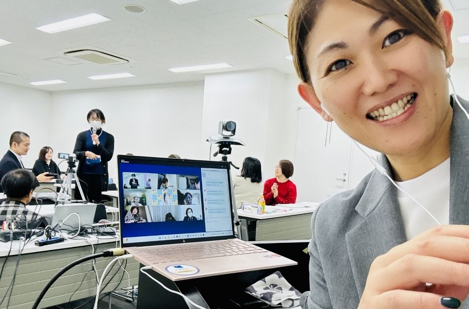 オンライン会議ツールの画面が映るノートパソコンを前に、イヤホンマイクをつけて笑顔で自撮りをする進絵美さんの写真。背景にはセミナー会場の参加者の姿も見える。