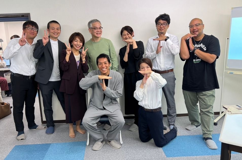 研修室のような場所で笑顔で集合写真に納まる山梨義正氏と関係者たち。参加者の数名が手で「T」の字を作るポーズをとっている。