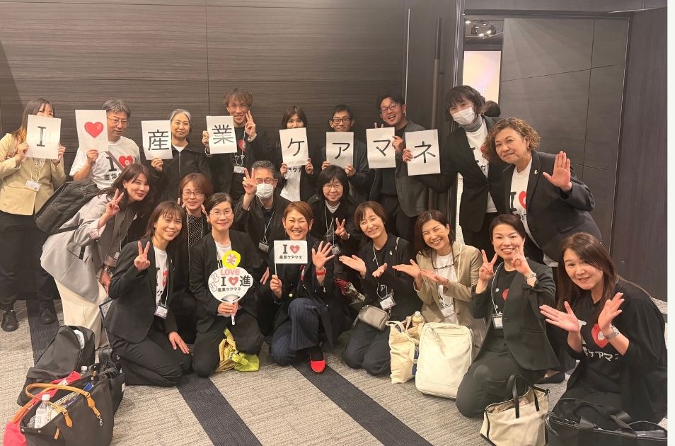 『I LOVE 産業ケアマネ』と書かれたTシャツを着たり、文字が書かれた紙を持ったりしている大勢の仲間たちに囲まれ、中央で笑顔を見せる進絵美さんの集合写真。