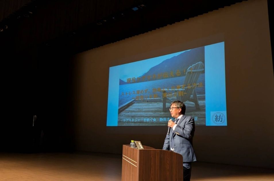 マイクを持って壇上で講演する山梨義正氏。背景の大きなプロジェクター画面には「現役ケアマネが伝える! ストレス溜めず、効率よく仕事する方法」のスライドが表示されており、「ケアマネジャーを紡ぐ会 静岡支部」のロゴが見える。