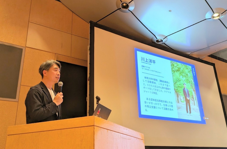 広いホールのステージに立ち、マイクを握って講演を行う川上洋平さんの写真。背後のスクリーンには、体育大学卒業や運動指導員を経てケアマネジャー10年目といった自己紹介が映し出されている。
