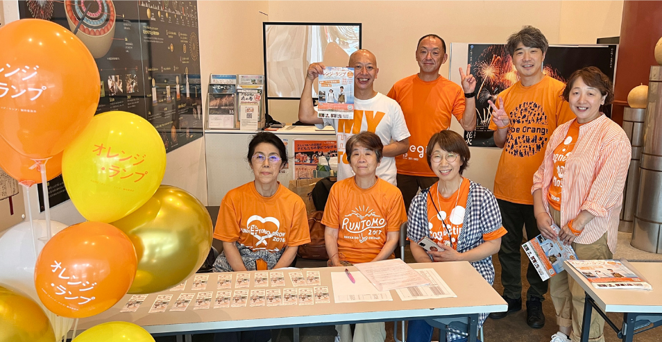 映画『オレンジ・ランプ』のロゴが入ったオレンジ色のTシャツを着たスタッフたちと、風船やポスターに囲まれて笑顔で写真に納まる川上洋平さんの集合写真。