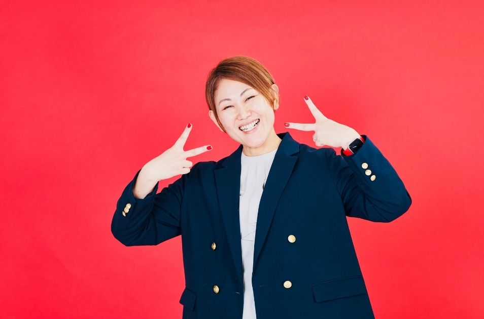 真っ赤な背景の前で、満面の笑みを浮かべて両手でピースサイン(ダブルピース)をする進絵美さんの写真。