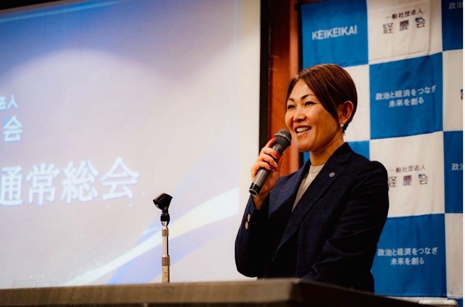 『一般社団法人 経慶会 通常総会』と書かれた看板を背景に、マイクを持って笑顔でスピーチをする進絵美さんの写真。