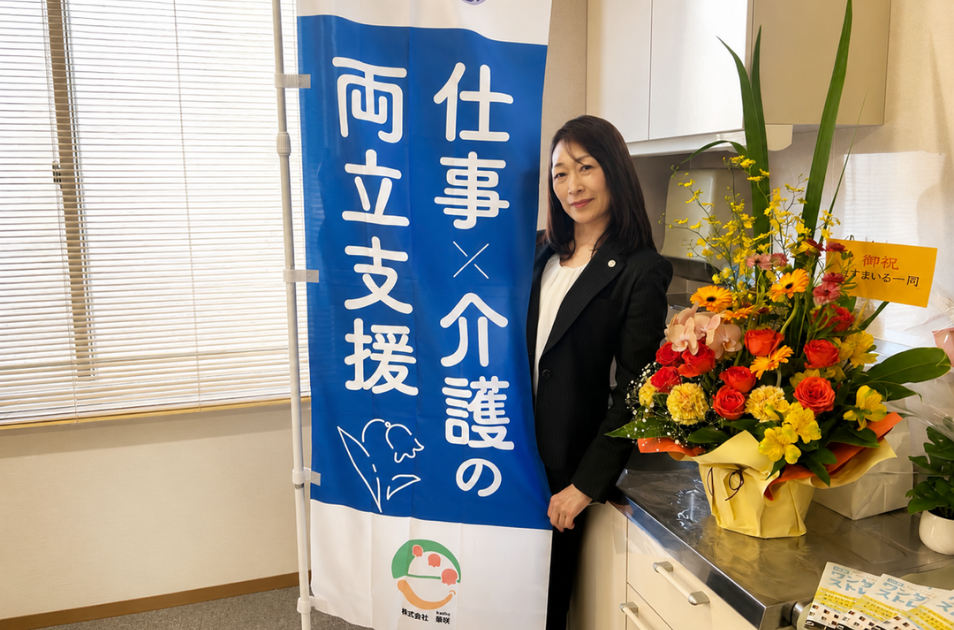 『仕事×介護の両立支援 株式会社華咲』と書かれた青いのぼり旗と、華やかなお祝いの花に囲まれて笑顔で立つ古池さんの写真。