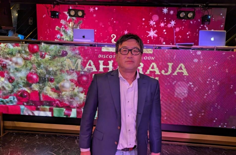 マハラジャ(MAHARAJA)のステージ前に立つジャケット姿の山梨義正氏。背景のスクリーンにはクリスマスの装飾が映し出されている。