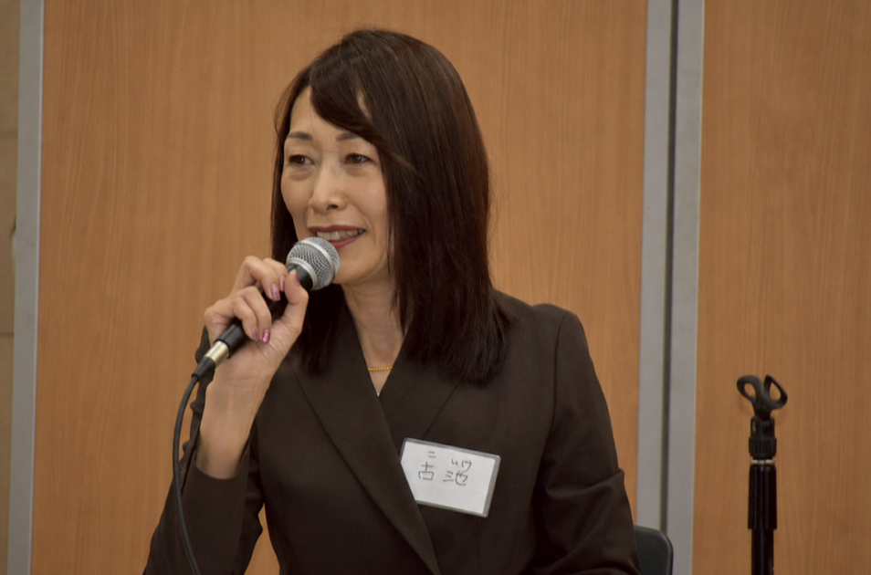 『古池』と書かれた名札を胸につけ、マイクを握って穏やかな笑顔でスピーチをしている古池さんのバストアップ写真。
