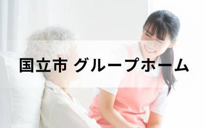 介護コラム：国立市のグループホーム事情。ピンクのエプロンを着た若い介護スタッフが、笑顔で高齢者に語りかけている安心感のあるイメージ。
