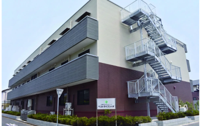 白とダークブラウンを基調とした3階建てのモダンな建物の外観完成予想図。手前には『介護付有料老人ホーム ベストライフつくば』と書かれた看板が立っており、建物の側面には外階段が設置されている。