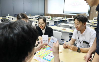 広い会議室で、数人の男女がテーブルを囲んで研修用のボードゲームを行っている写真。参加者は手にイラストや文字が書かれたカードを持ち、意見を交わしながらゲームを進めている。