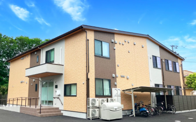 青空を背景にした、2階建ての認知症対応型共同生活介護施設『愛の家グループホーム所沢下安松』の外観写真。ベージュ、白、茶色を組み合わせた外壁で、エントランスには車椅子用のスロープが設置されている。