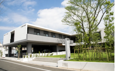 青空とシンボルツリーの緑を背景に建つ、白とグレーを基調としたモダンな2階建ての介護施設『特別養護老人ホーム 上白根の郷』の外観写真。