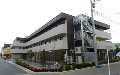 住宅街の角地に建つ、ダークブラウンと白を基調とした3階建ての介護施設「エクラシア足立舎人」の全体が写った外観写真。