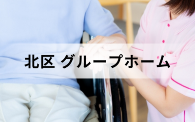 東京都北区のグループホーム。ピンクのユニフォームを着た介護スタッフが、車椅子に乗った高齢者の手を優しく握っている安心感のあるイメージ。