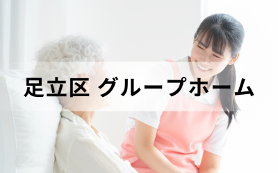 東京都足立区のグループホーム。ピンクのエプロンを着た若い介護スタッフが、笑顔で高齢者に語りかけている安心感のあるイメージ。