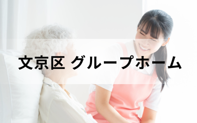 介護コラム：文京区のグループホーム事情。ピンクのエプロンを着た若い介護スタッフが、笑顔で高齢者に語りかけている安心感のあるイメージ。