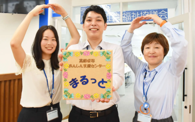花柄の装飾で『高齢者等あんしん支援センター まるっと』と書かれた看板を笑顔で持ち、頭の上で両腕を使って『まる』のポーズをとっている社会福祉協議会のスタッフ3名の写真。