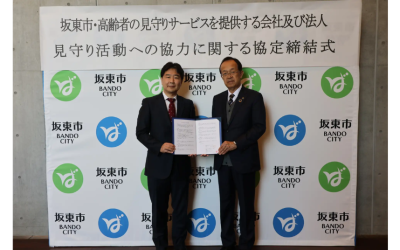 「坂東市・高齢者の見守りサービスを提供する会社及び法人 見守り活動への協力に関する協定締結式」の看板と坂東市のロゴが並ぶ背景幕の前で、スーツ姿の2人の男性が締結した協定書を開いてカメラに向けている記念写真。