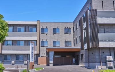 青空を背景にした、3階建ての介護付有料老人ホーム『イリーゼ新潟中央 アクティブ・リビング』の外観写真。グレー系のタイル調で統一されたモダンな建物で、手前には駐車場とエントランスの車寄せが設けられている。