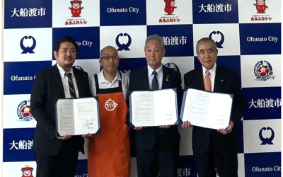 大船渡市のロゴやマスコットキャラクターが描かれたバックボードの前で、連携協定書を手に持ち、並んで笑顔で写真に収まる大船渡市の関係者と事業者の4名。