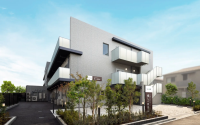 青空を背景に建つ、介護施設『癒しの新宿北』の外観写真。薄いグレーのタイル張りにガラス張りのバルコニーを備えたモダンな3階建ての建物で、エントランス前には緑豊かな植栽と焦げ茶色の施設看板が配置されている。