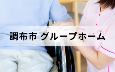 東京都調布市のグループホーム。ピンクのユニフォームを着た介護スタッフが、車椅子に乗った高齢者の手を優しく握っている安心感のあるイメージ。