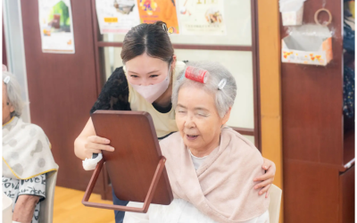 介護施設で、マスクとエプロン姿の女性スタッフが、頭にカーラーを巻いた笑顔の高齢女性に鏡を見せている写真。